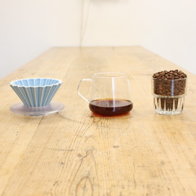 Links ein mattblauer Origami Dripper, in der Mitte ein Kinto Kännchen, gefüllt mit Filterkaffee. rechts Filterkaffeebohnen in einem Glas. Alles steht auf einem Holztisch.